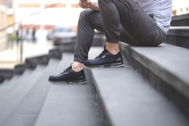 A person is sitting on steep, gray steps. They are wearing black shoes and comfortable, gray pants. The background shows blurry parts of buildings and people. The atmosphere seems relaxed.