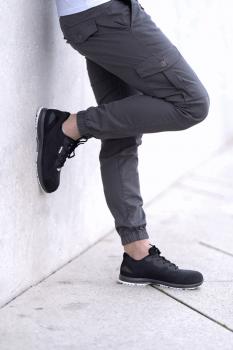 One leg is leaning against a wall, the wearer is dressed in gray cargo pants and black sneakers. The pose appears casual and relaxed. The background is light and solid-colored.