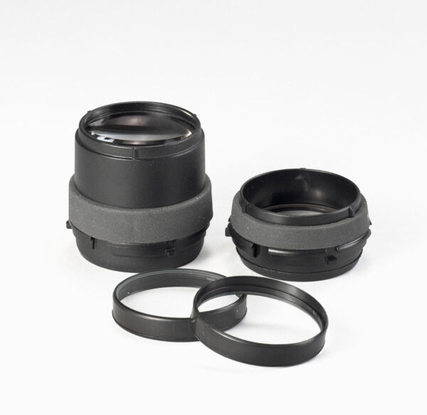 The image shows two black camera lenses and two black rings. The lenses have a round shape and are partially surrounded by a gray material. They are placed on a white surface.