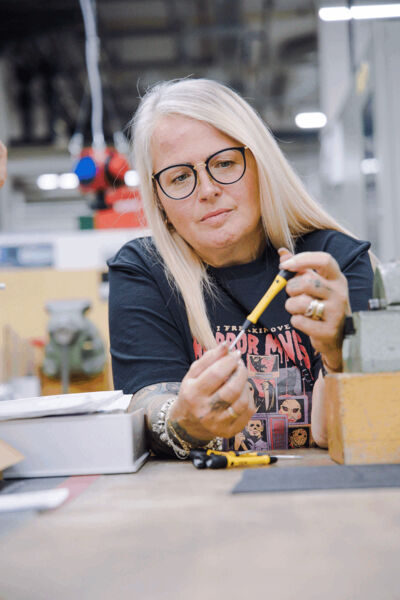 Eine lächelnde Frau mit langen, blonden Haaren und Brille hält einen gelben Bleistift. Sie sitzt an einem Tisch mit Werkzeugen und plastikartigen Objekten, umgeben von einer Werkstattatmosphäre.