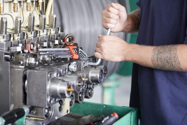 A worker in a blue shirt is repairing a machine component. He is holding a wrench and working on a metal block with many pipes and connections.