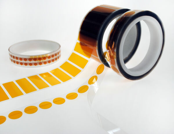 The image shows several rolls of tape. A large, orange roll with a transparent backing stands upright, while a strip with orange dots and a narrow, transparent tape lie next to it.