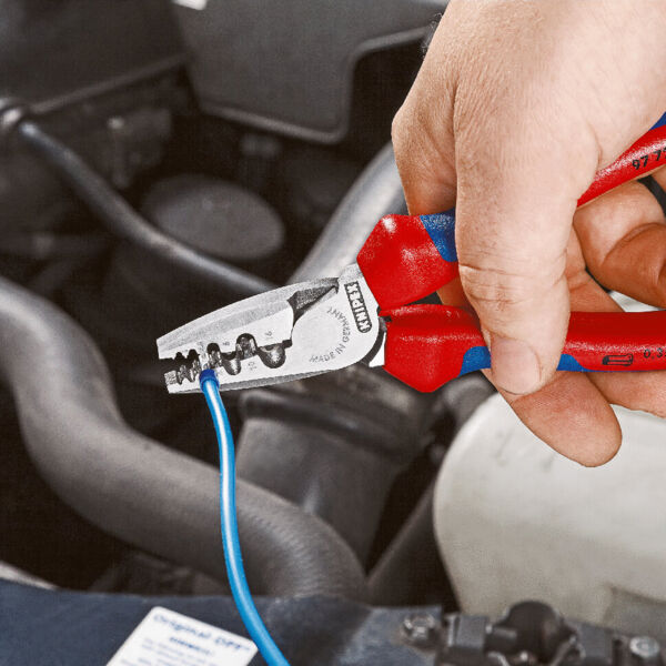 A hand holds red pliers that are gripping a blue wire. In the background, auto parts and hoses are visible. The pliers have a metallic blade and a non-slip grip area.