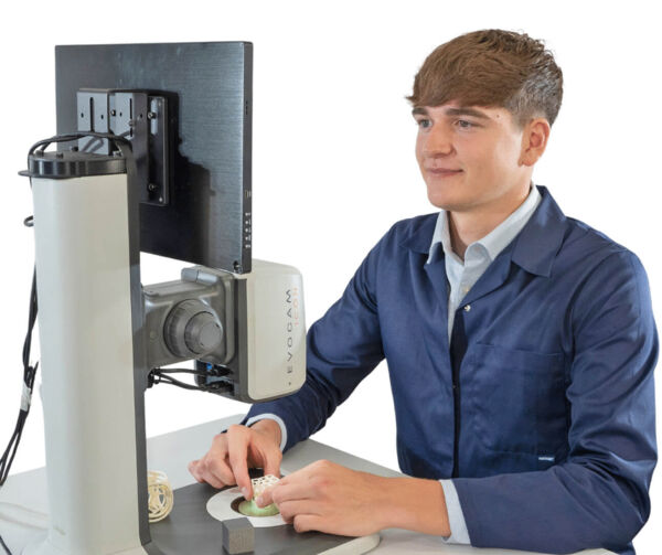 A young man is sitting at a table, in front of him a monitor and a magnifying device. He is looking intently at a small object that he is holding with one hand.