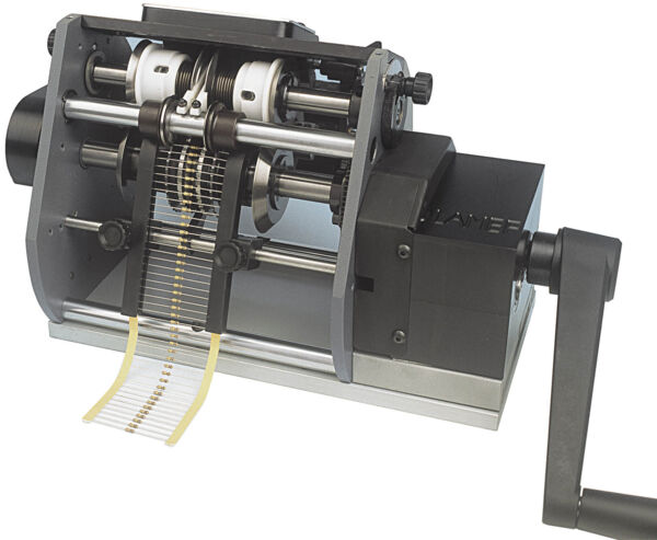 The image shows a mechanical machine with visible gears and rollers. A hand crank is located on the right side, and a long, thin strip of paper extends from the machine.