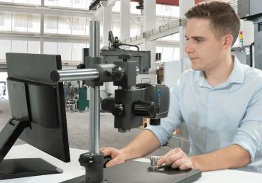 A young man is sitting at a table in front of a monitor. In front of him is a microscope with a large lens, mounted on a sturdy stand. He is focusing on his work.