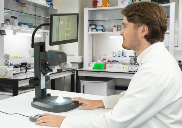 A man in a lab coat is sitting at a table, looking at a monitor connected to a microscope. In the background, shelves with laboratory utensils are visible.