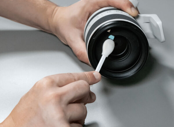 A hand holds the lens ring of a camera. The lens is being cleaned with a cotton swab. The background is plain and inconspicuous.