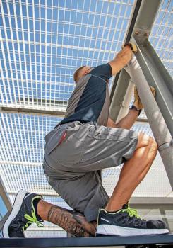 Ein Mann klettert auf eine Struktur. Er trägt Shorts und ein kurzärmliges Shirt. Seine Füße stecken in sportlichen Schuhen. Der Himmel ist blau mit einem Gitter über ihm.