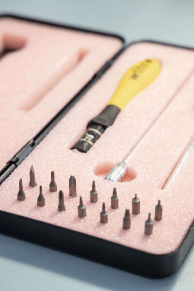 The image shows a screwdriver with a yellow handle, surrounded by various metallic inserts in a pink foam padding. There are a total of 16 bits available.