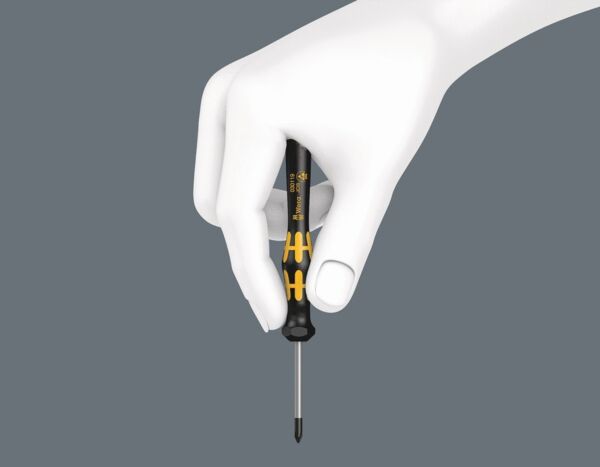 The image shows a hand holding a screwdriver with a black handle and yellow accents. The screwdriver has a pointed blade and is held vertically. The background is gray.