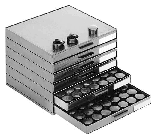 The image shows a stackable box with multiple drawers. Some drawers are open and contain round, flat objects. The top is shiny and has two lockable containers.