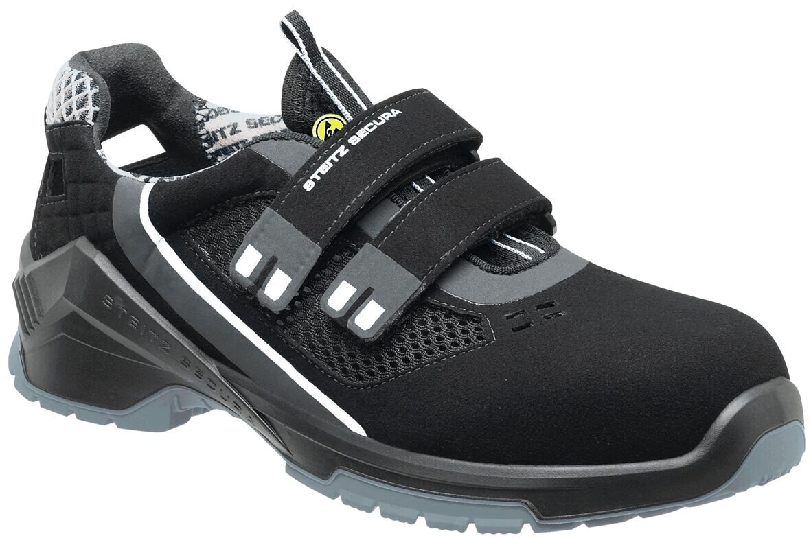 The image shows a black, sporty safety shoe with two wide Velcro straps. The shoe has a textured sole and breathable elements. It appears sturdy and functional.