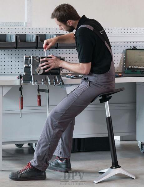 A mechanic is sitting on an adjustable stool and working on a machine. He is wearing gray work clothes and using tools that are lying on a table.