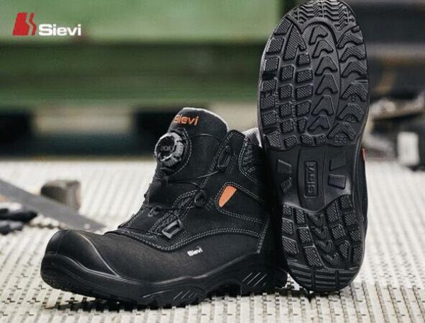 The picture shows a black work boot with a rugged treaded sole. The boot reaches up to mid-ankle, features a quick-lacing system, and has an orange detail.