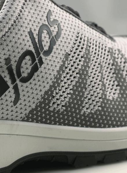 The image shows a sporty shoe in gray and white with a modern, textured upper. The brand name is printed on the side in black. The sole is dark and non-slip.