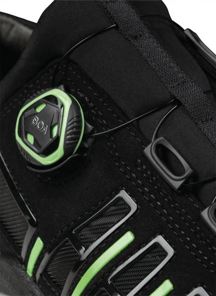 The image shows a part of a sporty shoe in black with green accents. A round closure mechanism is prominent to adjust the lacing. There are also side slits.