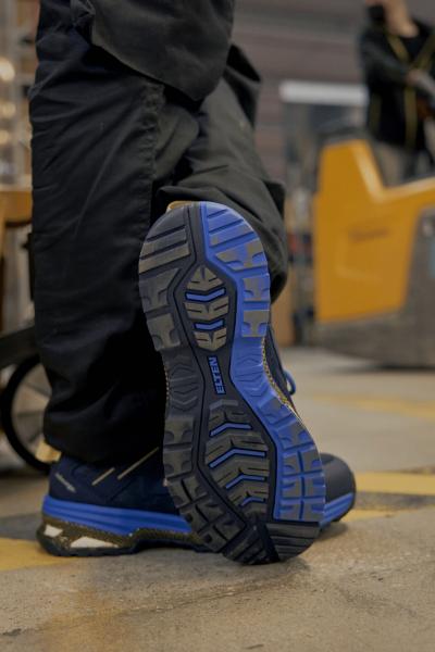 The image shows the underside of a blue shoe on a floor. The sole features a distinctive, non-slip tread with various patterns and lines that provide good traction.