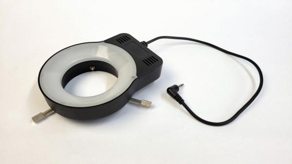 The image shows a round light ring with a black casing, which is connected on one side by a cable. The interior is bright, and the ring has two side mounts.