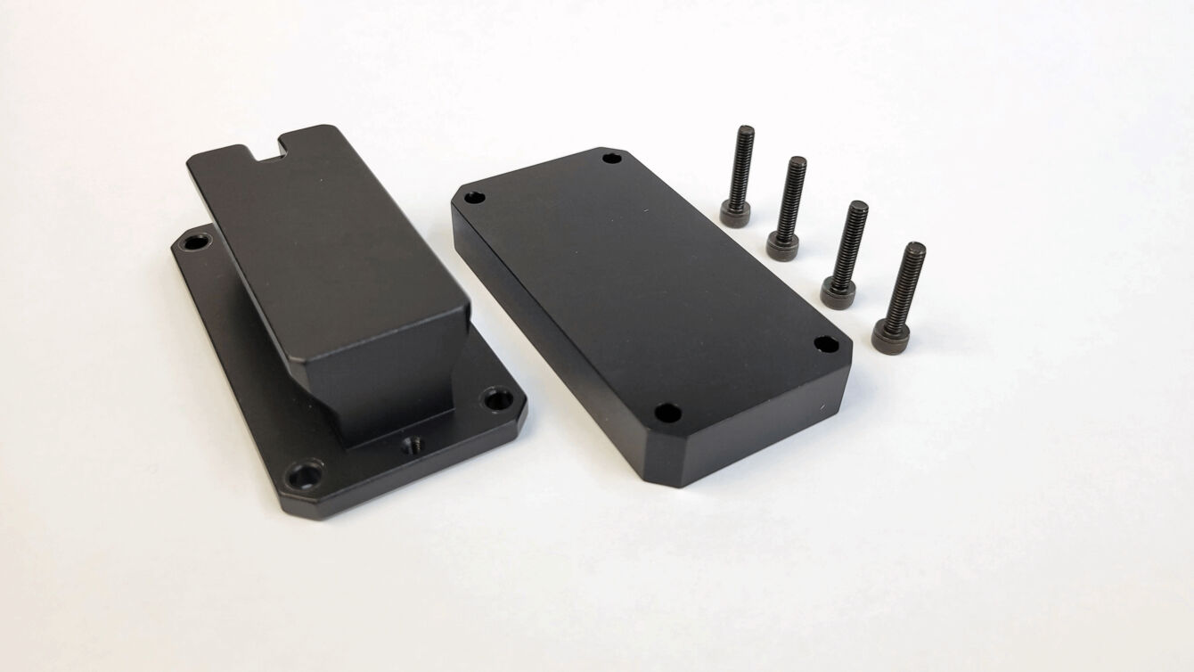 The image shows two black metal parts in rectangular shape. One has a small elevation, the other is flat. There are also four black screws. The background is white.