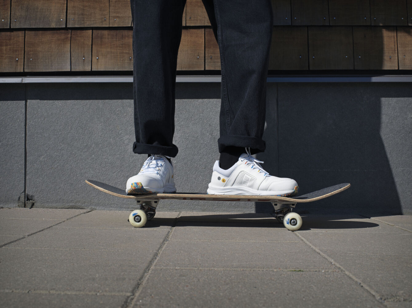 In the picture, a person is standing on a skateboard. They are wearing white sneakers and dark, baggy pants. The background consists of wooden panels and a gray wall.