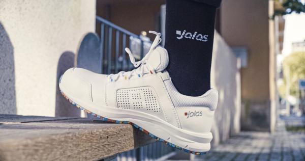 The image shows a white sports shoe standing on a wooden board. A black sock with a white logo is visible. In the background, stairs and buildings are suggested.