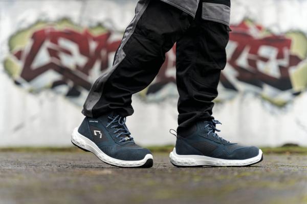 In the image, there are simple, sporty shoes in dark blue with white soles. The person is wearing black, loose pants. In the background, there is a graffiti-like, colorful wall print.