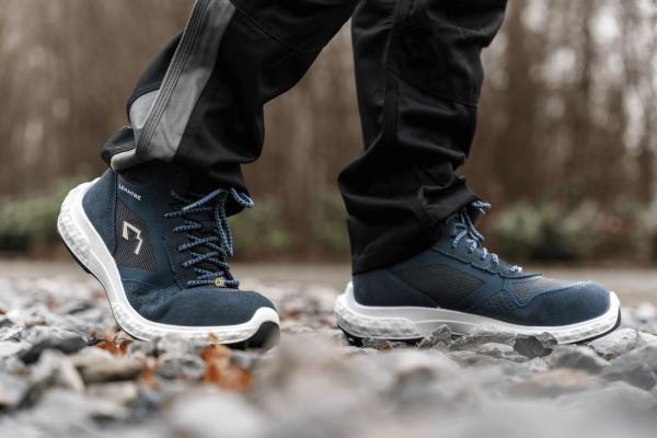 The image shows a person standing on a rocky surface. They are wearing dark blue, sporty shoes with light soles and are dressed in dark pants. The background is blurred.