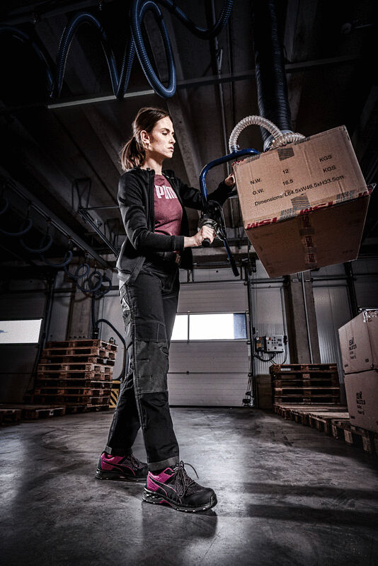 A woman is lifting a box in a warehouse. She is wearing work clothes and safety shoes. In the background, wooden pallets and a modern warehouse environment are visible.