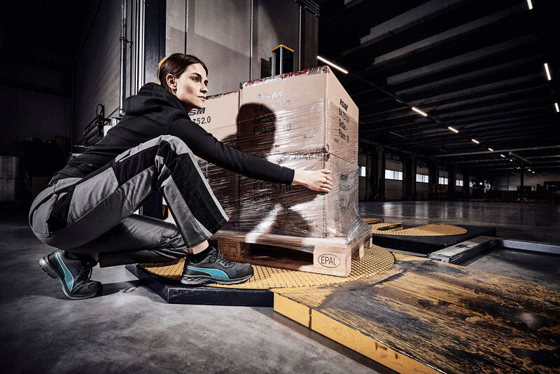 A person in athletic clothing bends down to secure a box on a pallet. The background shows a warehouse with large windows and industrial lighting.