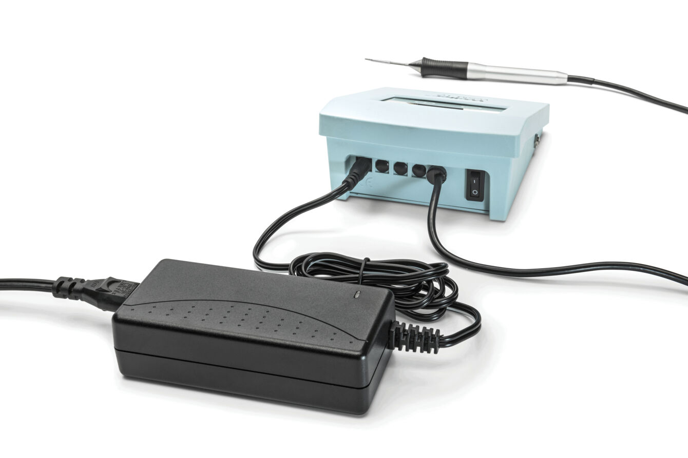 The image shows a device with a light blue box that has cable connections. Next to it is a black power supply with a long cable. A soldering iron is also lying next to it.