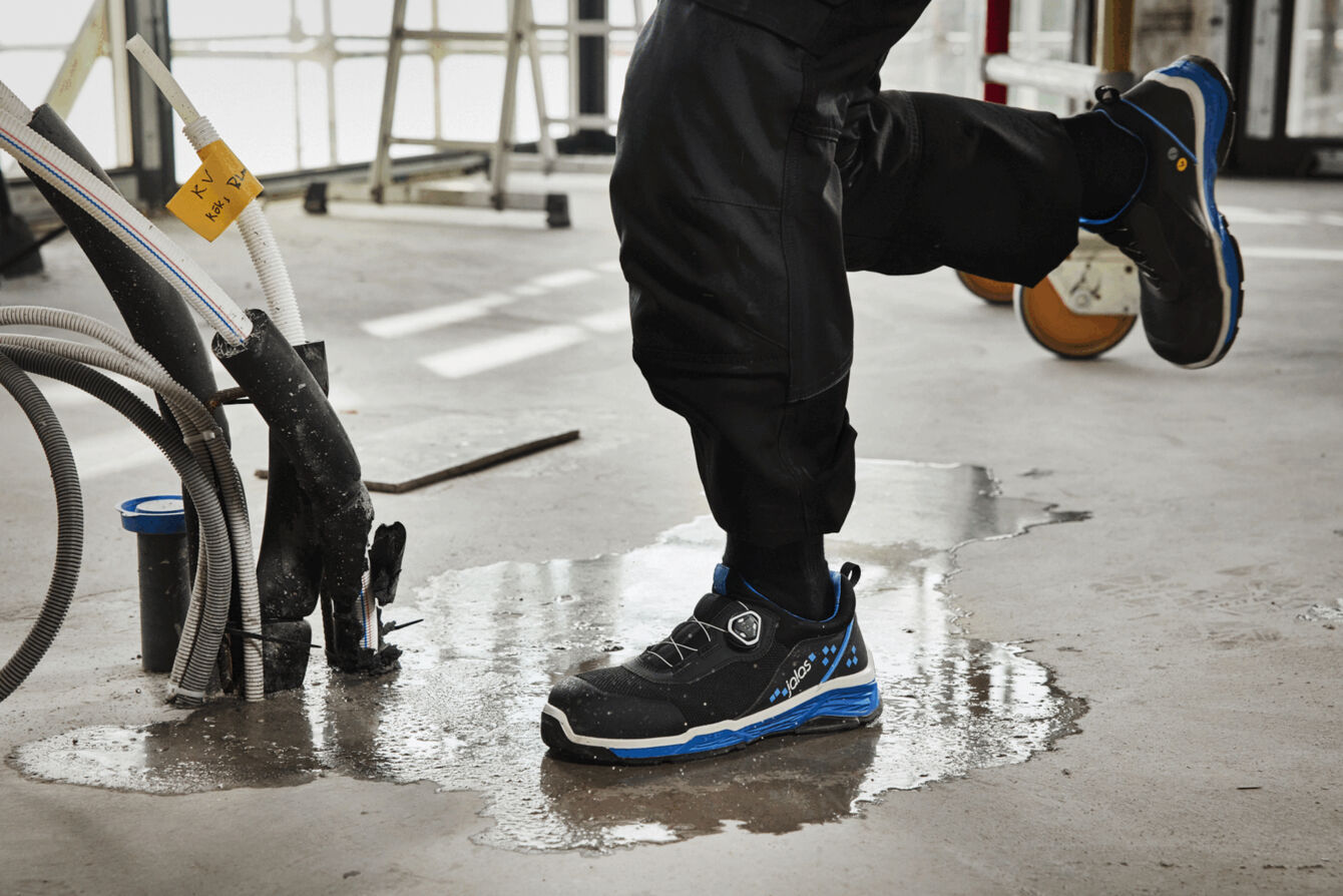 A foot stands on a wet floor, surrounded by tools and hoses. The shoe is black with blue accents, the pants are dark and protect against moisture.