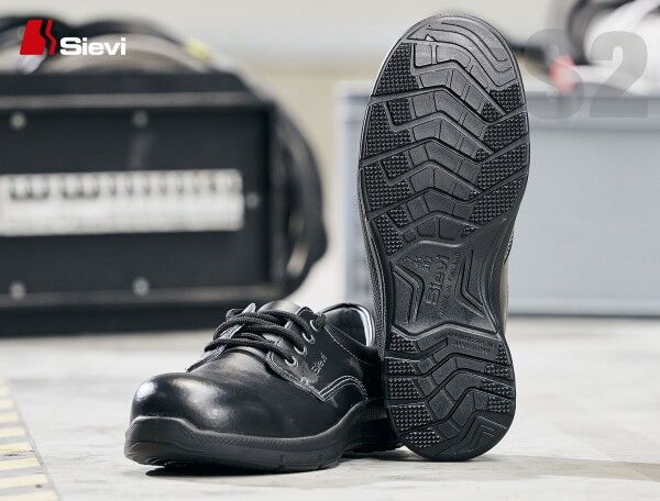 The image shows a black safety shoe from Sievi. The shoe features simple lacing and a textured, non-slip sole with a distinct tread. In the background, tools are blurred.