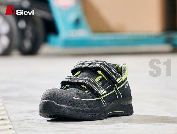 The image shows a black safety shoe with yellow accents and two Velcro straps. It has a sturdy sole and is designed for occupational safety. Backgrounds are blurred.