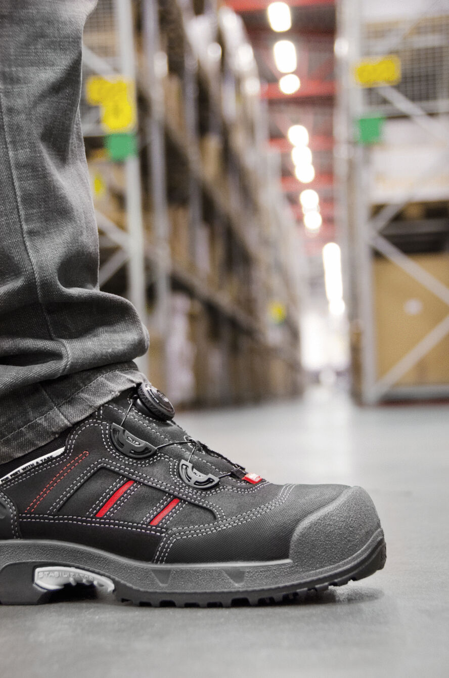 The image shows a foot wearing a black safety shoe with red accents. In the background, shelves with boxes and a wide warehouse can be seen.