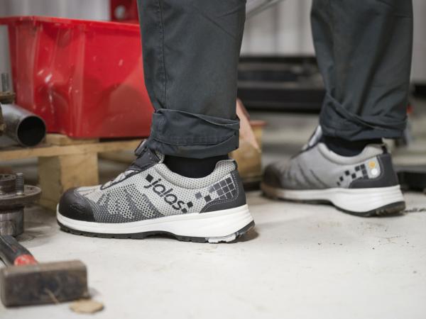 In the picture, there are two feet in sturdy, gray work shoes standing on a neutral floor. Around them, some tools and a red box are visible.