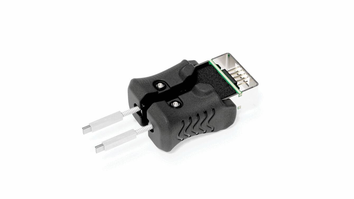 The image shows a black plug with a rectangular connection on top and two metallic pins protruding from the sides. The surface is ribbed and there are small screws.