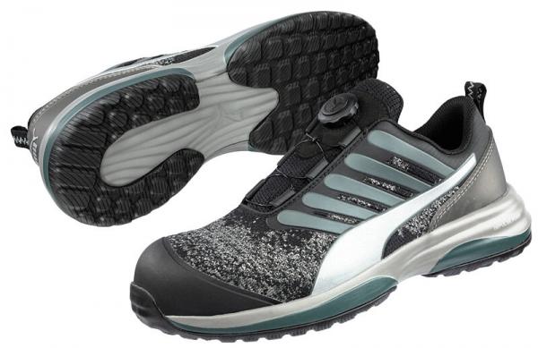 The image displays a modern sports shoe in black and gray with a textured sole. The shoe features a quick lacing system and a padded, stable outer sole for good grip.