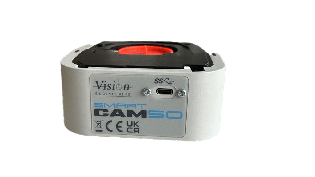 The image shows a compact, rectangular camera in white with a black lid. On the front, there is a round red button and a connection visible. The bottom has a label.