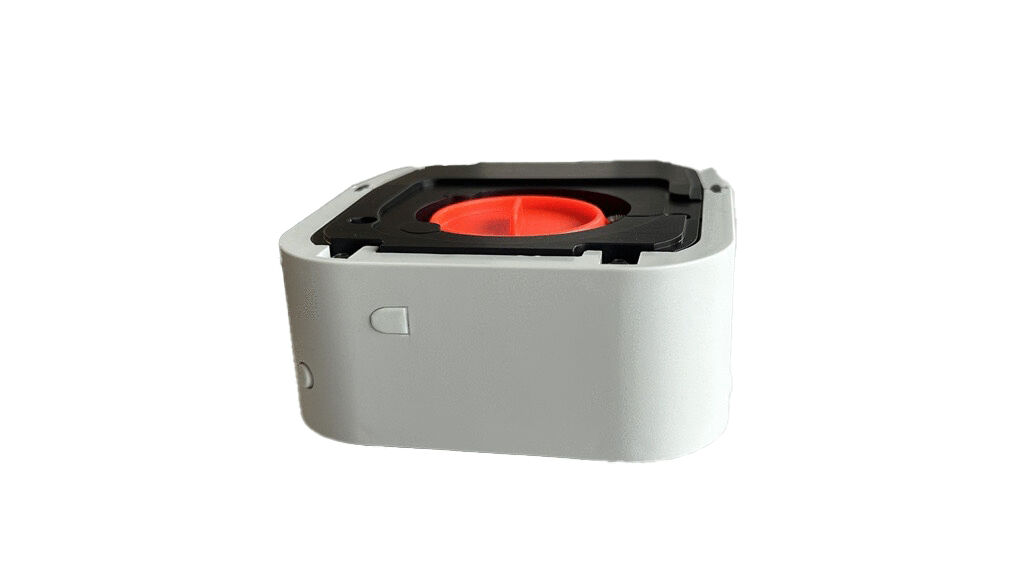 The image shows a compact, cuboid device in white with a red, round opening in the center. The top has a dark, rectangular structure that frames the red part.