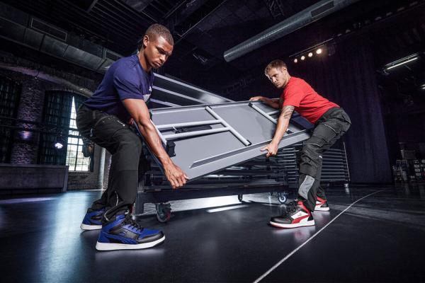 Two men are lifting a table together in a modern room. One is wearing a blue shirt and sneakers, the other a red shirt and black pants. They are working intently.