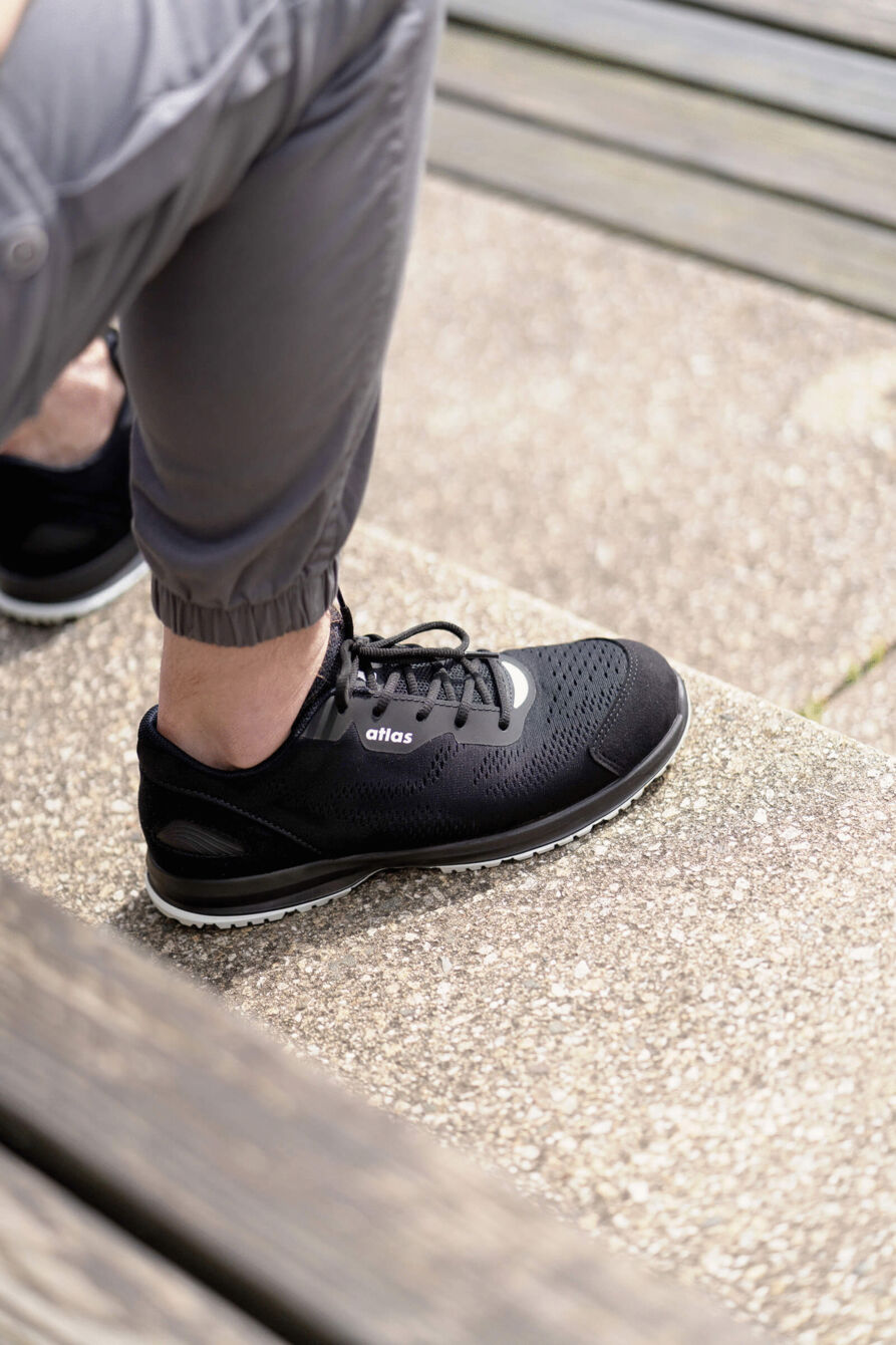 In the picture, there is a foot in a black athletic shoe. The shoe has a smooth surface and a light-colored sole. The foot is wearing a gray, elastic pants. In the background, wooden planks can be seen.