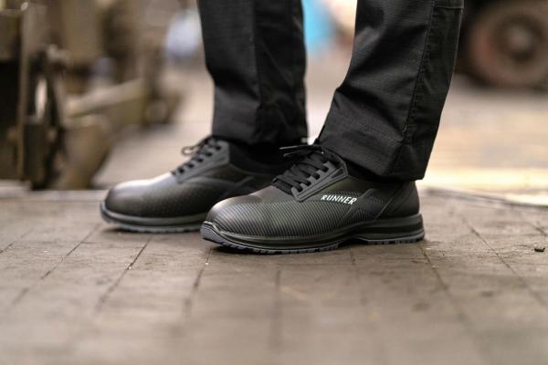 In the picture, there are black work shoes with a textured surface visible. The shoes are laced and are on a wooden or asphalt floor. A person is wearing dark pants.