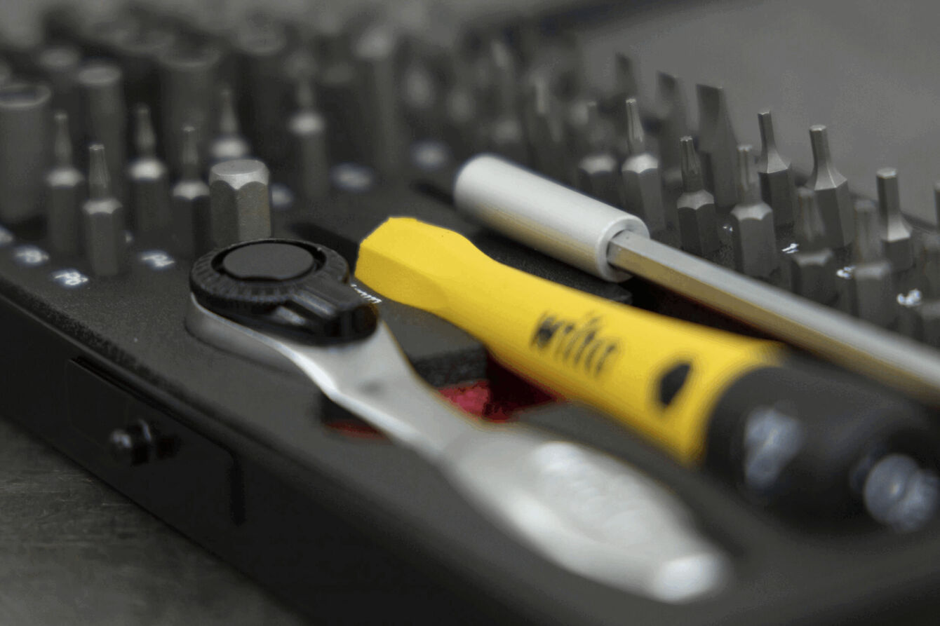 The image shows a toolset with various bits and a ratchet. In the center lies a yellow screwdriver. The tools are arranged on a dark background.