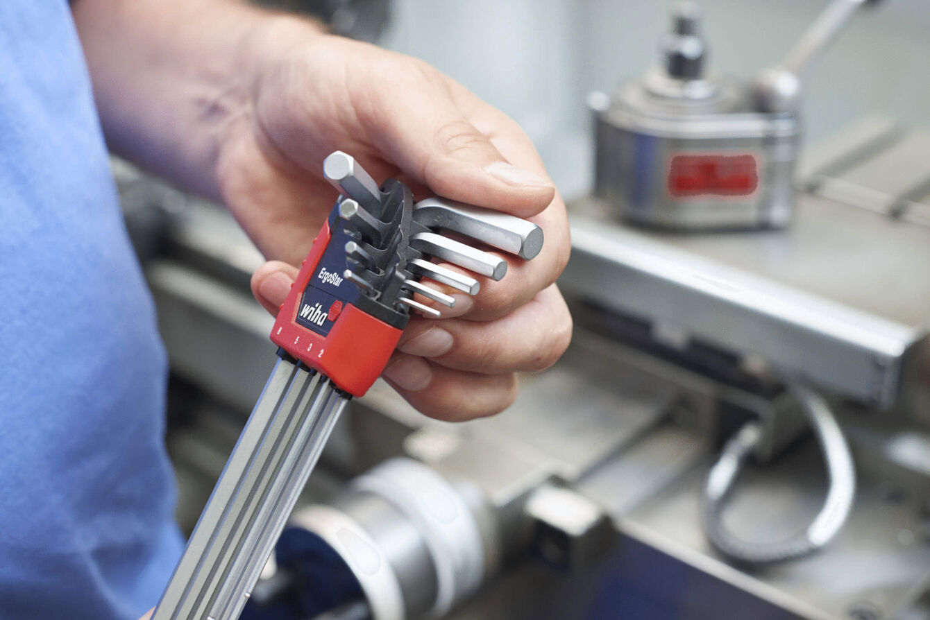 A hand holds a set of hex keys. The keys are in various sizes and arranged frontally. In the background, machines and tools are visible.