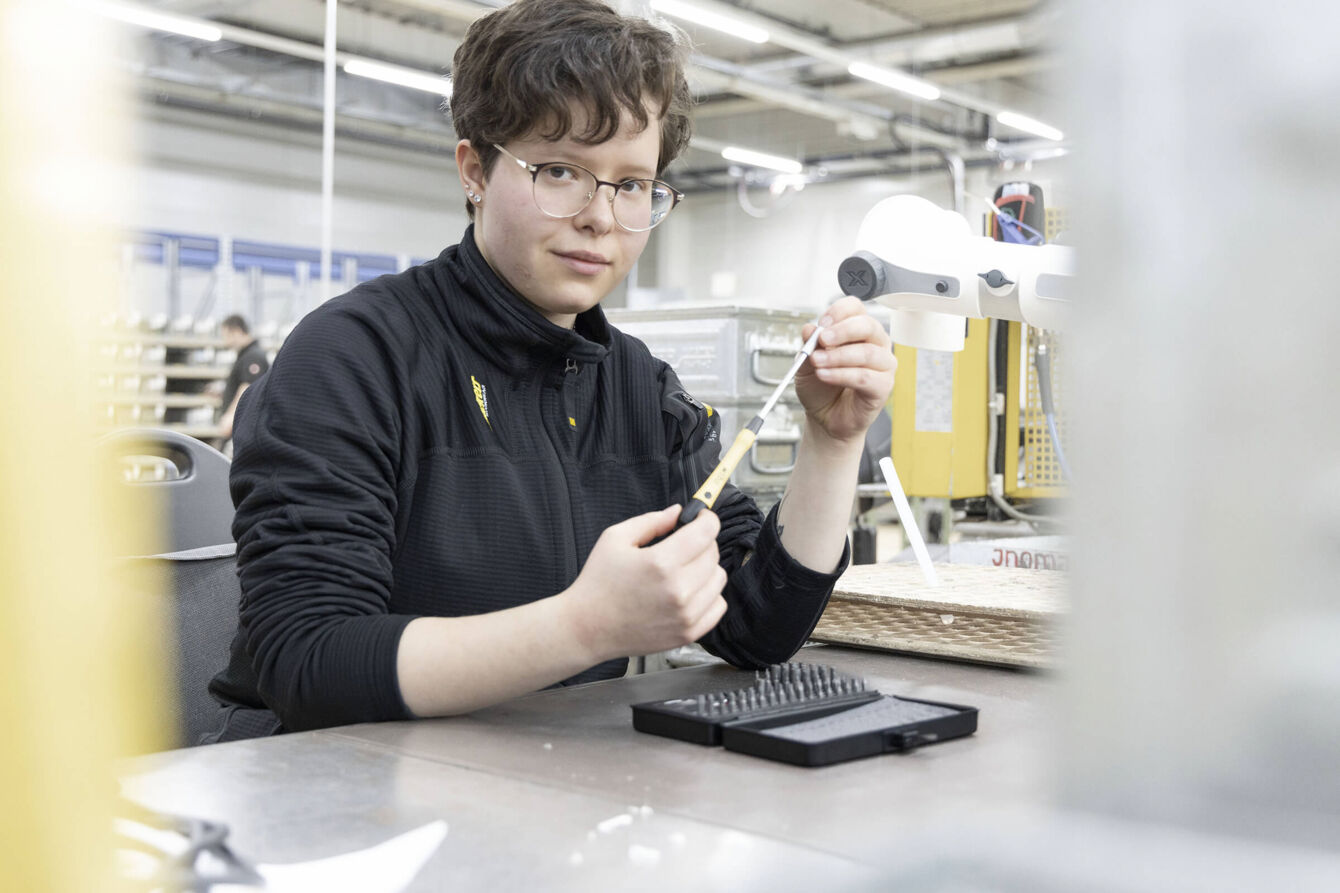 A person with short, curly hair is sitting at a table in a workshop. They are wearing a black jacket and holding a tool in their hand while looking intently at the camera.