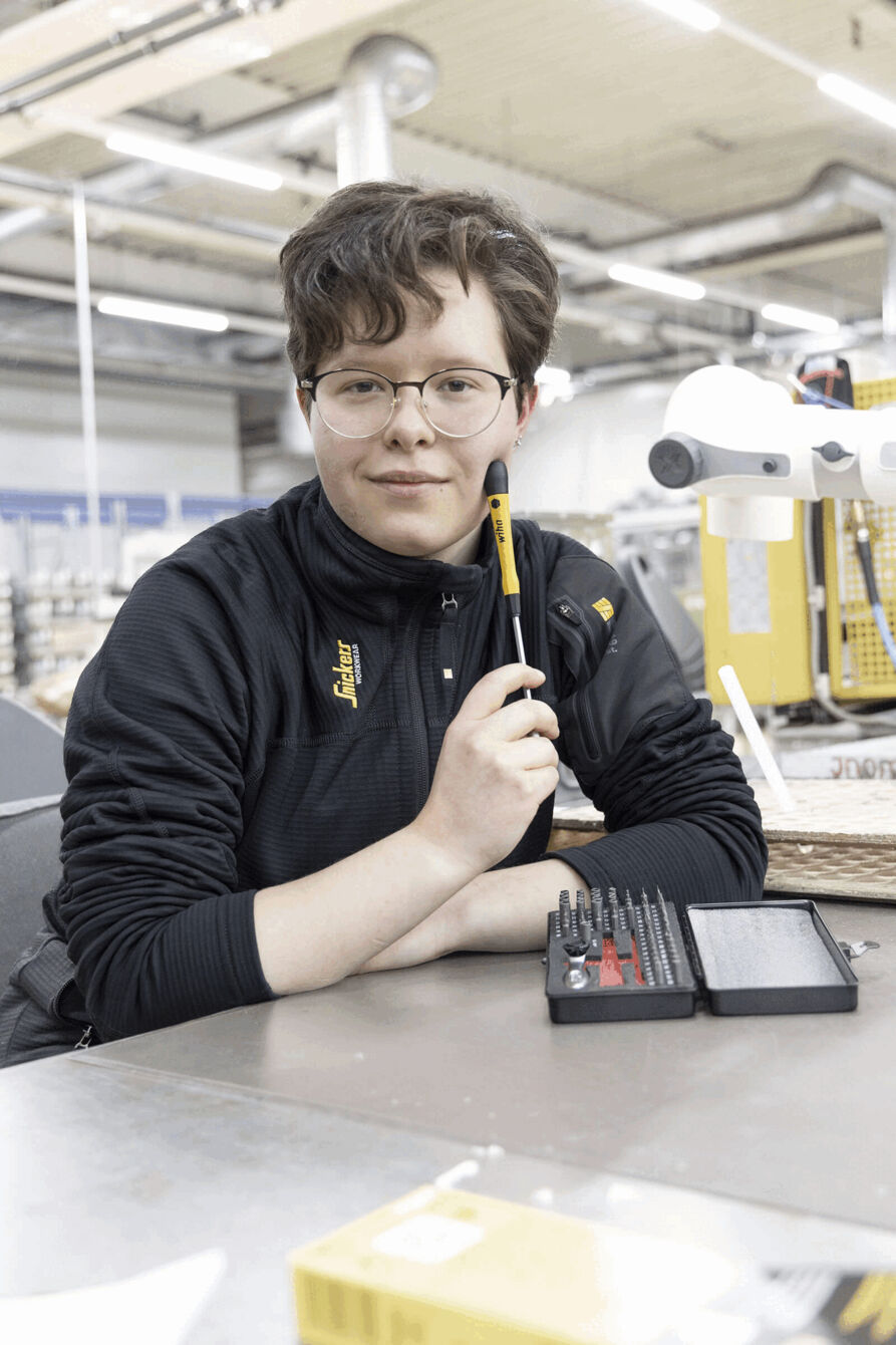 A person with curly, brown hair and glasses is sitting at a table. They are holding a pencil in their hand, and in front of them lies a toolbox with various screwdrivers.