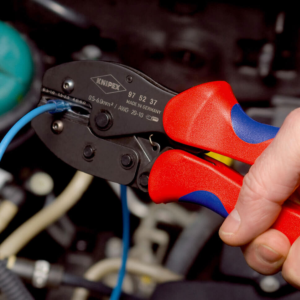 The image shows a hand holding a red and black cable clamp. A blue wire is being worked on with it. In the background, cables and parts of an engine are visible.