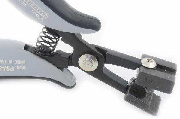 The image shows a black pair of pliers with gray handles and a spring. It features a special blade at one end and a set screw, which likely serves for adjustment.