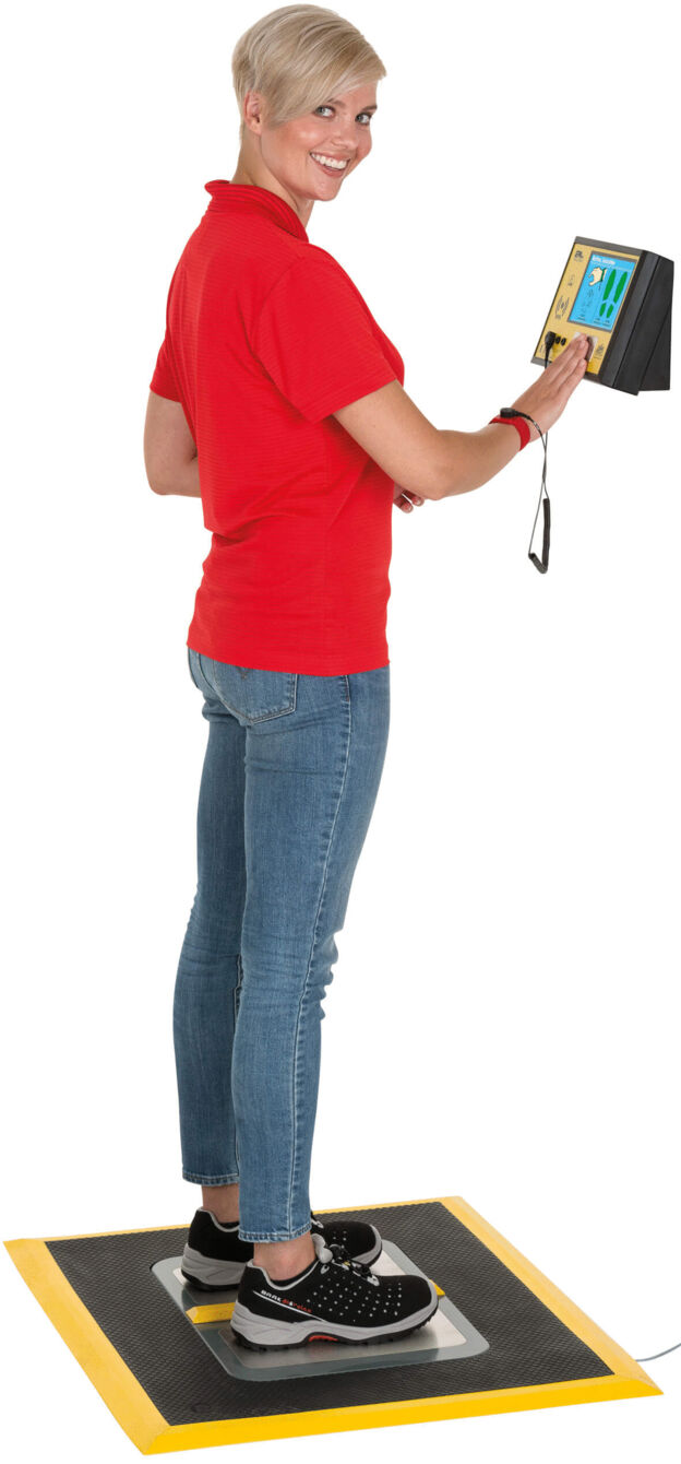 Eine Person mit kurzen, blonden Haaren trägt ein rotes Shirt und Jeans. Sie steht auf einer Plattform und hält ein digitales Gerät in der Hand. Der Hintergrund ist neutral.